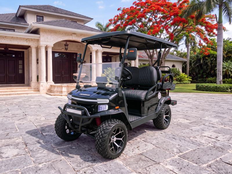 Denago Nomad XL lifted four-seat electric golf cart in black on a grey stone forecourt in front of a coral stone villa with mahogany doors and a red flamboyant tree in bloom in Casa de Campo