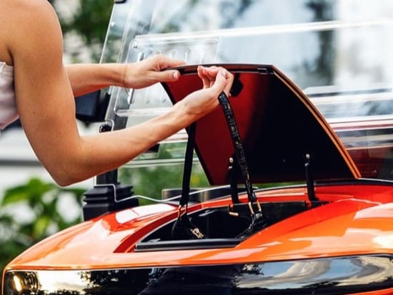 Hand lifting the lid of the storage compartment under the front hood of an orange Denago golf cart