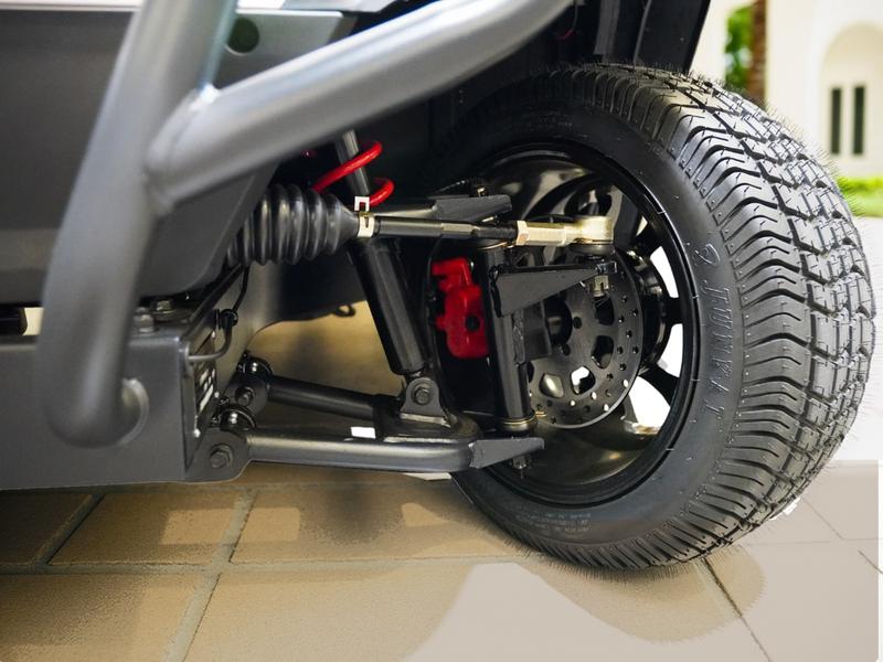 Front suspension of a Denago golf cart with double wishbone arms, a red coil spring, steering rack and a red disc brake caliper, on a villa driveway in Casa de Campo