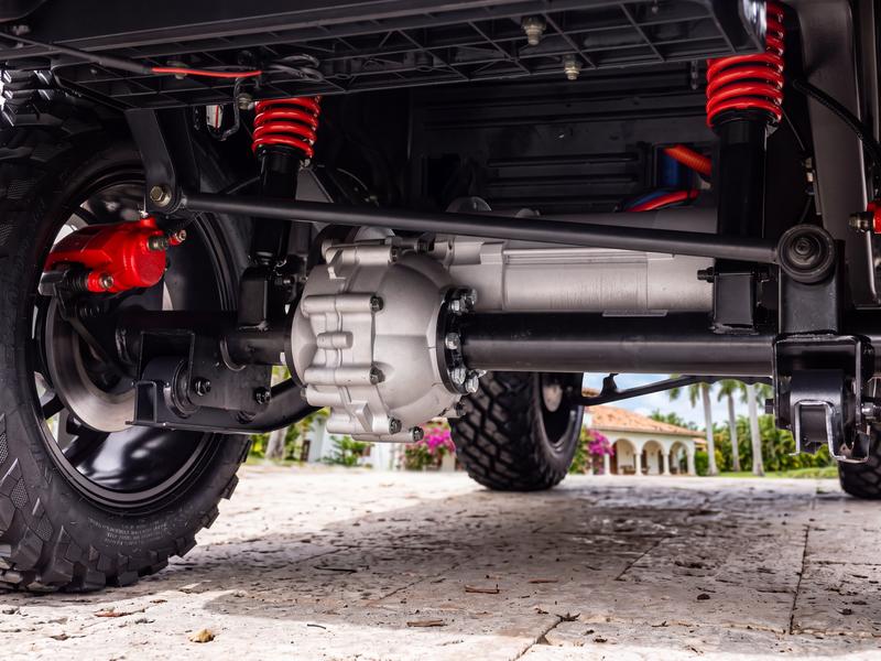 Rear axle of a Denago golf cart seen from below, with the electric motor and gearbox, red coil-over shocks and a red disc brake caliper, on a villa driveway in Casa de Campo
