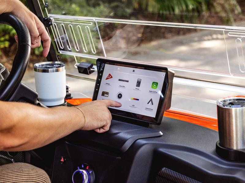 Driver tapping the 10.1 inch touchscreen of a Denago golf cart, with Apple CarPlay and Android Auto on the home screen
